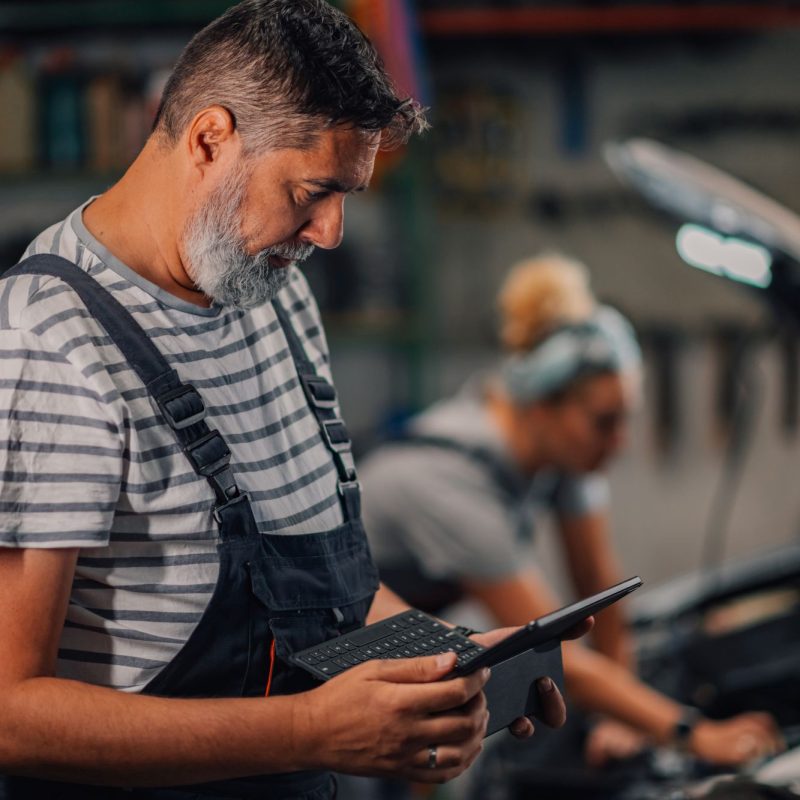 Side view of auto mechanic doing car diagnostics on laptop at wo Profile view of male auto mechanic worker standing at workshop with little laptop in hands and doing car diagnostics while looking at it. In a blurry background is female mechanic fixing a car at shop